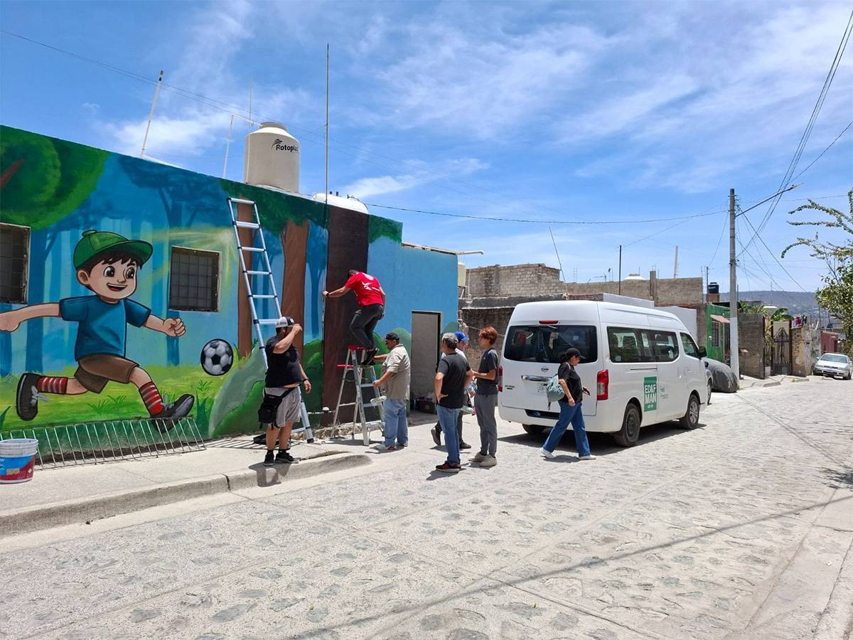 Mural of a smiling boy kicking a soccer ball on a blue wall; several people and a ladder nearby on a sunny street.